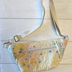 May include: A quilted fanny pack featuring a patchwork design with yellow, orange, and blue floral and plaid patterns. The bag includes a horizontal zippered front pocket, a silver-toned metal zipper pull, and an adjustable cream-colored fabric strap.