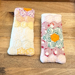 May include: Two rectangular quilted cases for eyeglasses, constructed from patchwork fabric in a hexagonal pattern. The left case features yellow, orange, and white floral prints. The right case displays a central orange hexagon surrounded by red, white, and green floral and geometric patterns.