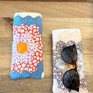 Puede incluir: Dos estuches rectangulares de tela acolchada con un diseño de patchwork hexagonal. Uno muestra patrones florales en rojo, blanco y azul con un centro naranja. El otro presenta un patrón floral en negro, blanco y melocotón con gafas de sol oscuras.