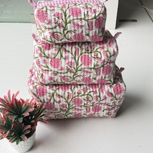 Op de afbeelding: Een set van drie gestapelde rechthoekige gewatteerde tasjes met een witte basis en een patroon van roze bloemen en groene ranken. Elk tasje heeft een roze-witte vichy-ruitjesrand aan de bovenkant en kleine stoffen lipjes aan de zijkanten.