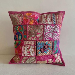 Puede incluir: Un cojín decorativo cuadrado con un diseño de patchwork. El cojín presenta una variedad de cuadrados de tela bordada en tonos de rosa, dorado y azul. Los bordes están ribeteados con una tela rosa lisa.