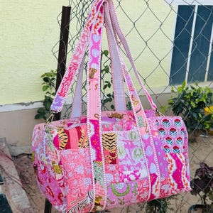 May include: A rectangular duffel bag featuring a patchwork design in shades of pink, magenta, and white. The bag displays various patterns including floral motifs, tiger stripes, and geometric shapes, with two long fabric handles attached to the top.