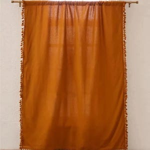 May include: A single, burnt orange curtain hangs from a gold-colored rod in front of a window. The curtain is trimmed with small, matching tassels along the edges. The fabric is slightly sheer, allowing some light to pass through.