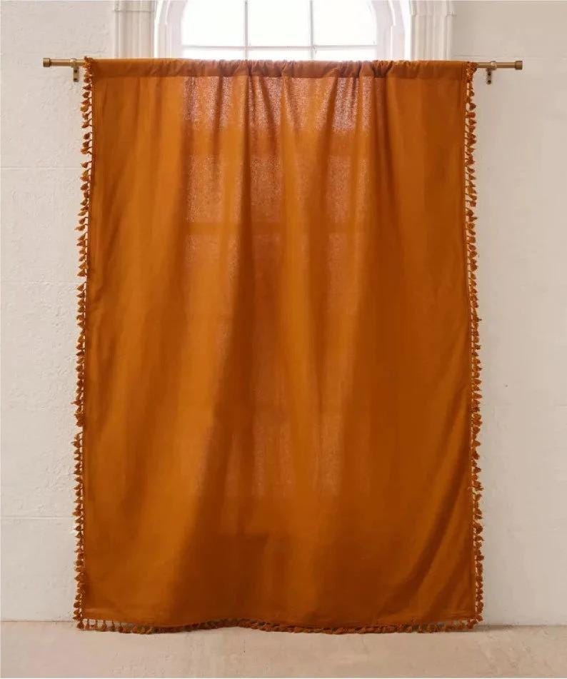 May include: A single, burnt orange curtain hangs from a gold-colored rod in front of a window. The curtain is trimmed with small, matching tassels along the edges. The fabric is slightly sheer, allowing some light to pass through.