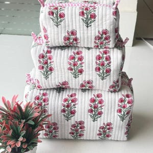 Peut inclure: Une pile de trois trousses rectangulaires matelassées en tissu blanc avec un motif floral imprimé en rose et vert. Chaque trousse présente une bordure à carreaux roses et blancs sur le dessus et de petites languettes en tissu sur les côtés.