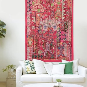 Puede incluir: Un tapiz de patchwork rosa y rojo con bordados intrincados cuelga de una pared blanca sobre un sofá blanco con almohadas verdes y blancas.