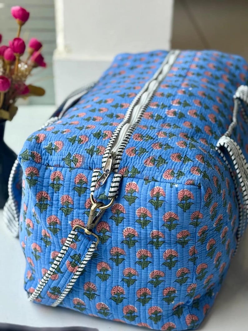 Puede incluir: Una bolsa de viaje acolchada azul con un estampado floral en rosa y verde. La bolsa tiene una cremallera blanca y correas a rayas blancas y negras. La bolsa est&aacute; hecha de una tela acolchada suave y tiene forma rectangular.