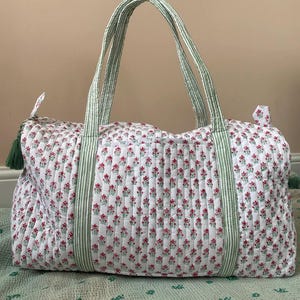 Puede incluir: Una bolsa de lona acolchada blanca con un estampado floral de peque&ntilde;as flores rojas y hojas verdes. La bolsa tiene asas y detalles a rayas verdes y blancas. Una borla verde cuelga de la cremallera.