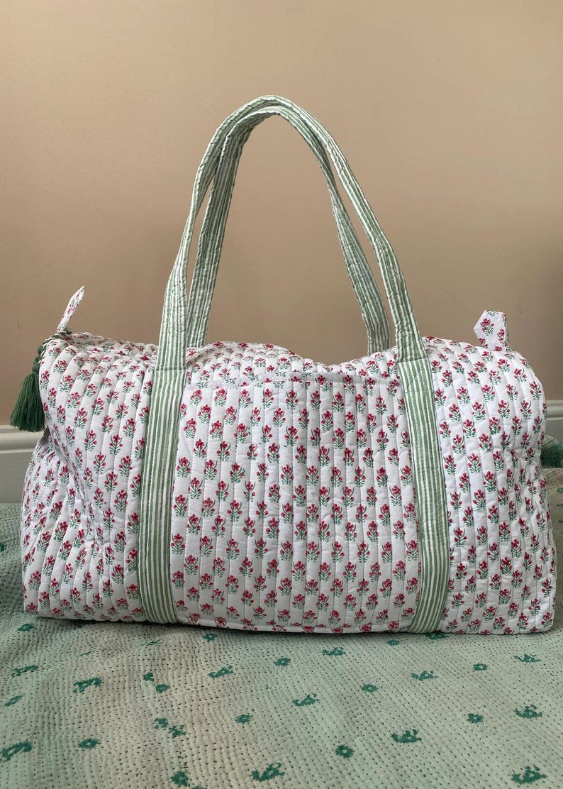 Puede incluir: Una bolsa de lona acolchada blanca con un estampado floral de peque&ntilde;as flores rojas y hojas verdes. La bolsa tiene asas y detalles a rayas verdes y blancas. Una borla verde cuelga de la cremallera.