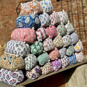 Puede incluir: Una pirámide de pequeñas bolsas rectangulares de tela con varios patrones florales y geométricos en colores como naranja, azul, rosa y verde. Cada bolsa tiene un pequeño lazo en la parte superior. Las bolsas están apiladas sobre una superficie de madera.