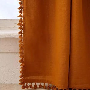 May include: Mustard-colored curtain with a textured appearance. The edges are trimmed with small, matching tassels. The curtain is hanging, with a glimpse of a white surface and window in the background.