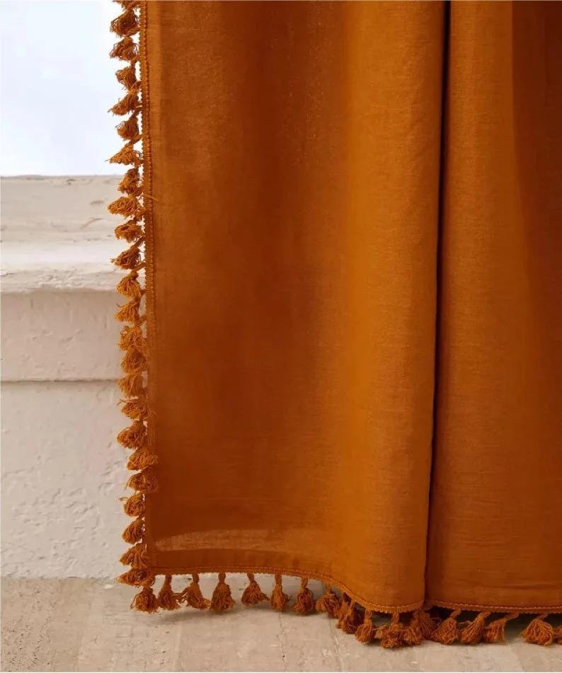 May include: Mustard-colored curtain with a textured appearance. The edges are trimmed with small, matching tassels. The curtain is hanging, with a glimpse of a white surface and window in the background.