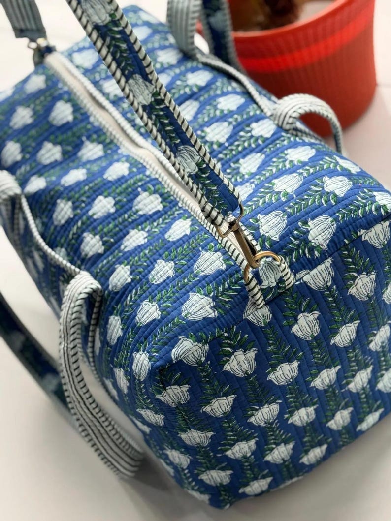 Puede incluir: Bolso de viaje azul con estampado floral blanco. Cuenta con una cremallera blanca y asas a juego. El dise&ntilde;o floral presenta flores blancas y hojas verdes sobre un fondo azul. Las asas son a rayas blancas y azules.