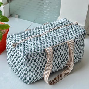 Könnte beinhalten: Eine rechteckige Reisetasche mit weißem und grünem Blumenmuster, Reißverschluss und beige gestreiften Griffen. Die Tasche ist aus gestepptem Stoff gefertigt und eignet sich zum Reisen oder Tragen von Gegenständen.