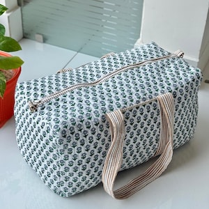 Puede incluir: Una bolsa de viaje rectangular con un estampado floral blanco y verde y cierre de cremallera. La bolsa tiene dos asas a rayas marrones y beige. La bolsa est&aacute; hecha de una tela suave y acolchada.