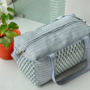 Puede incluir: Una bolsa de viaje rectangular con un estampado de rayas en blanco y azul marino. Los extremos de la bolsa presentan un estampado floral en blanco y azul. La bolsa tiene dos asas y un cierre de cremallera. Una peque&ntilde;a planta en una maceta naranja est&aacute; en el fondo.
