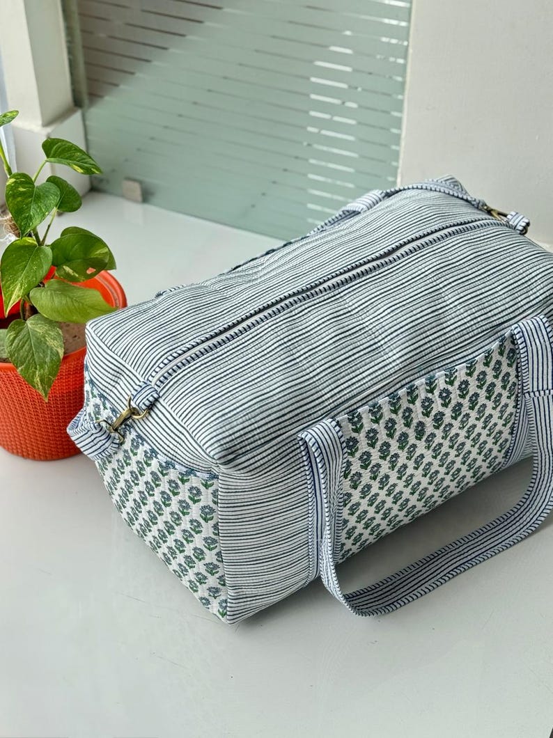 Puede incluir: Una bolsa de viaje rectangular con un estampado de rayas en blanco y azul marino. Los extremos de la bolsa presentan un estampado floral en blanco y azul. La bolsa tiene dos asas y un cierre de cremallera. Una peque&ntilde;a planta en una maceta naranja est&aacute; en el fondo.