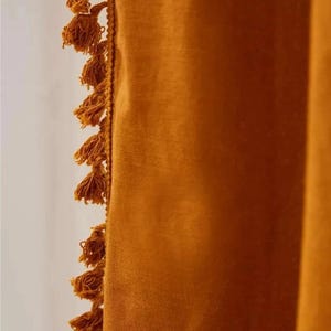 May include: Close-up of a mustard-colored curtain with a textured fabric. The edge is adorned with a row of small, matching tassels. The curtain's color is a warm, earthy tone, creating a cozy and inviting aesthetic. The tassels add a decorative element.