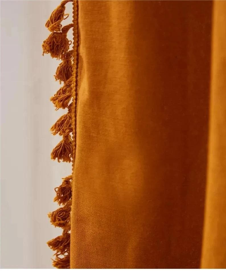 May include: Close-up of a mustard-colored curtain with a textured fabric. The edge is adorned with a row of small, matching tassels. The curtain's color is a warm, earthy tone, creating a cozy and inviting aesthetic. The tassels add a decorative element.
