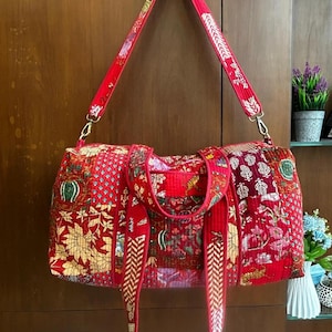 May include: A quilted duffel bag featuring a vibrant red patchwork design with various floral and animal patterns. The bag includes two short carrying handles and a long, adjustable shoulder strap, all attached with gold-toned metal hardware.