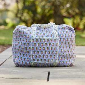 Puede incluir: Una bolsa de lona acolchada con un fondo azul claro y un patr&oacute;n repetido de fresas rojas con hojas verdes. La bolsa tiene forma rectangular, con un asa superior y un bolsillo frontal.