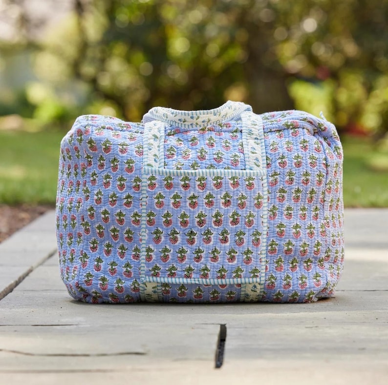 Puede incluir: Una bolsa de lona acolchada con un fondo azul claro y un patr&oacute;n repetido de fresas rojas con hojas verdes. La bolsa tiene forma rectangular, con un asa superior y un bolsillo frontal.