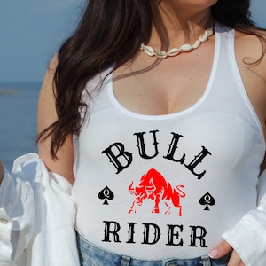 May include: White tank top with a black and red graphic of a bull and the text "BULL RIDER" with two black spade symbols.