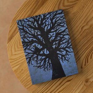 May include: A rectangular notebook with a painted illustration of a black tree with bare branches against a blue sky. A black cat with yellow eyes sits in the tree. The notebook is on a wooden surface.