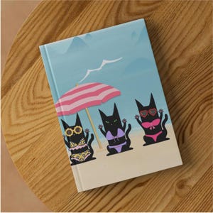 May include: A rectangular notebook featuring a light blue beach scene with three black cats wearing bikinis and sunglasses under a pink and white striped beach umbrella. The background shows a sandy beach, a pale blue sky, and distant mountains.