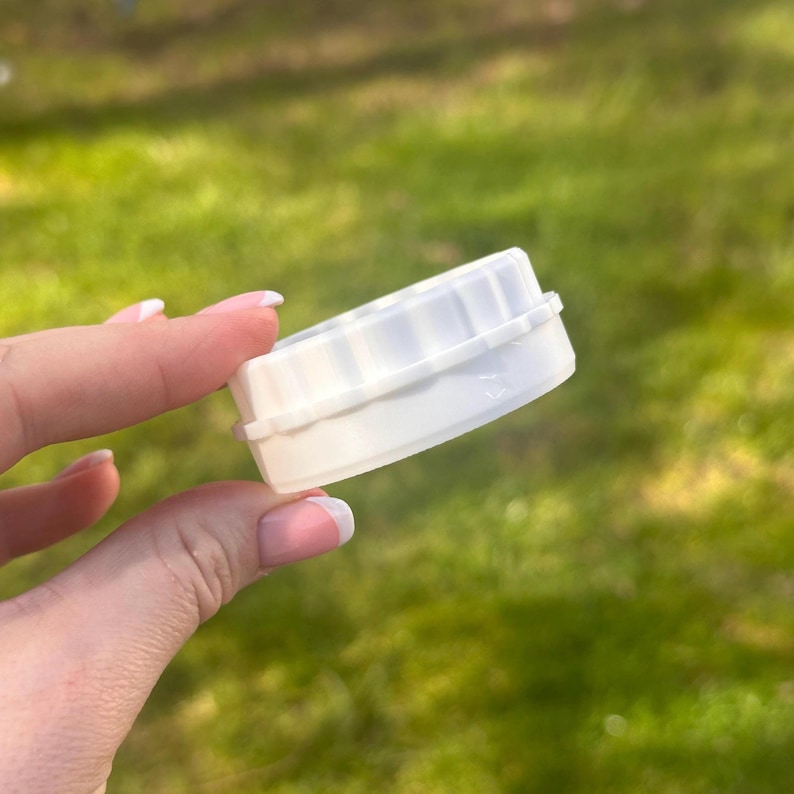 Op de afbeelding: Een witte plastic deksel met een geribbelde bovenkant en een groef aan de rand. De deksel wordt in de hand van een persoon gehouden tegen een vage groene achtergrond van gras.