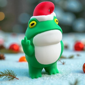 May include: A green frog figurine wearing a red and white Santa hat, giving the middle finger. The frog has yellow eyes and a white belly. Christmas ornaments and pine needles are in the snowy background.