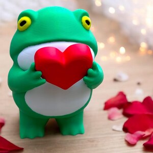 Può includere: Una statuetta di rana verde che tiene un cuore rosso. La rana è in piedi su una superficie di legno con petali di rosa rossa sparsi intorno.