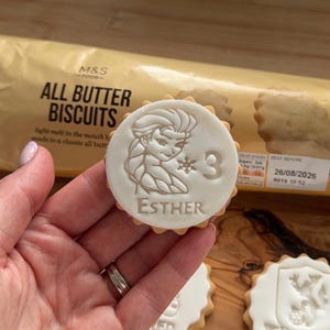 Op de afbeelding: Een ronde koek met een wit glazuurontwerp met een stripfiguur, het nummer 3 en de naam "ESTHER". De koek wordt in een hand gehouden, met een verpakking "ALL BUTTER BISCUITS" op de achtergrond.