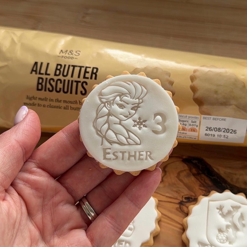 Op de afbeelding: Een ronde koek met een wit glazuurontwerp met een stripfiguur, het nummer 3 en de naam "ESTHER". De koek wordt in een hand gehouden, met een verpakking "ALL BUTTER BISCUITS" op de achtergrond.