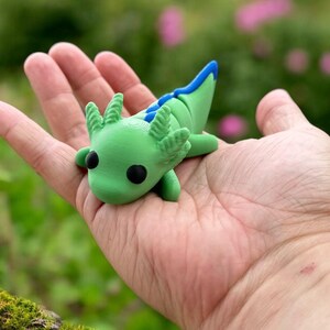 May include: A green and blue axolotl figurine with black eyes, sitting in the palm of a hand. The figurine is made of polymer clay and has a detailed design.