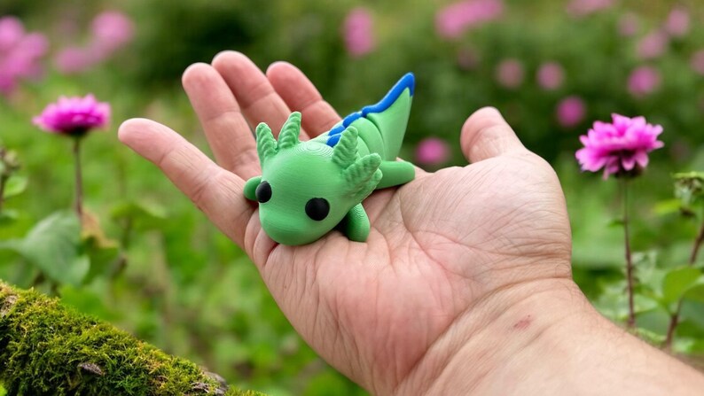 May include: A green and blue axolotl figurine with black eyes, sitting in the palm of a hand. The figurine is made of polymer clay and has a detailed design.