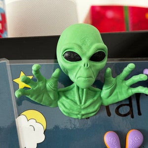 Op de afbeelding: Een groene alien-figuur met grote ogen en een serieuze uitdrukking. De alien heeft zijn armen uitgestrekt, met zijn handen in een grijpende positie. De figuur staat op een blauwe achtergrond met sterren en wolken.