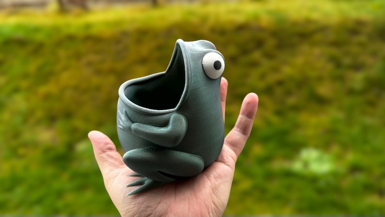 May include: A gray ceramic frog planter with a large open mouth and a single white eye. The frog is sitting on a hand.