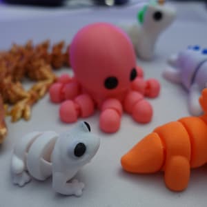 Puede incluir: Una colección de figuritas de animales articuladas, impresas en 3D y de colores vivos. Las figuritas incluyen un pulpo rosa, una rana blanca, un dinosaurio naranja, un dragón dorado y un unicornio blanco.