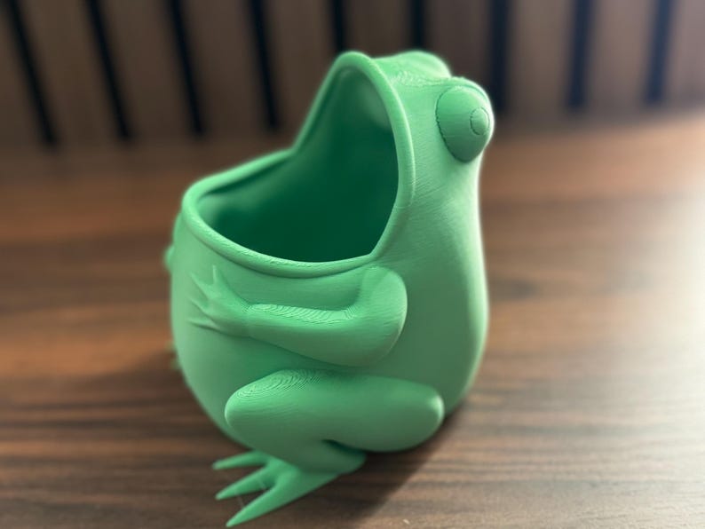 May include: A green frog-shaped planter with a wide opening at the top. The planter is made of a smooth, textured material and has a realistic frog design.