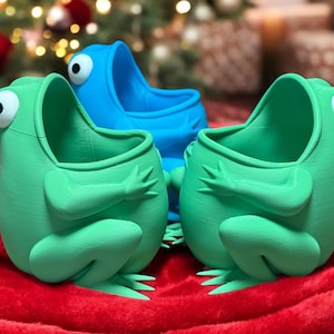 May include: Three quirky frog-shaped planters in green and blue, featuring large googly eyes and open mouths. The planters are set on a red blanket, with a Christmas tree in the background. These planters are ideal for small plants.