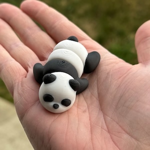 Puede incluir: Pequeña figura de panda en blanco y negro, tumbada boca arriba. El panda tiene un cuerpo redondeado con extremidades y orejas negras, y una cara blanca. La figura se sostiene en una mano abierta, mostrando su pequeño tamaño y diseño detallado.