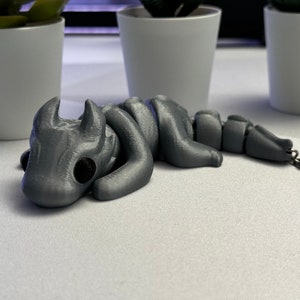 May include: A silver-coloured, segmented, articulated dragon keyring with black eyes. The dragon is curled up in a sleeping position.