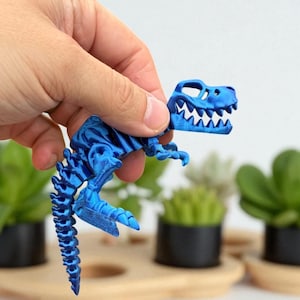 Puede incluir: Un juguete de esqueleto de dinosaurio azul articulado se sostiene en una mano. El juguete presenta un dise&ntilde;o detallado de un esqueleto de T-Rex, con costillas, columna vertebral y cr&aacute;neo visibles. El juguete mide aproximadamente 10 cm de largo.