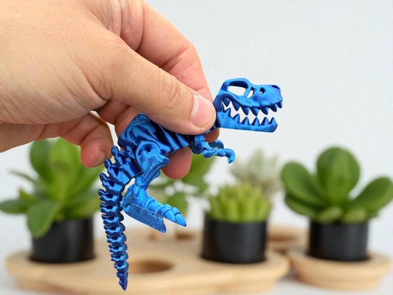 Puede incluir: Un juguete de esqueleto de dinosaurio azul articulado se sostiene en una mano. El juguete presenta un dise&ntilde;o detallado de un esqueleto de T-Rex, con costillas, columna vertebral y cr&aacute;neo visibles. El juguete mide aproximadamente 10 cm de largo.