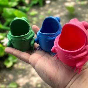 May include: Three frog-shaped planters, one green, one blue, and one pink. The planters are made of plastic and have a wide opening at the top.