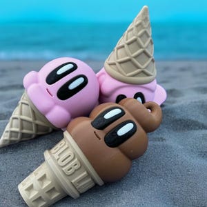 May include: Two ice cream cone shaped toys on a sandy beach. One is pink with black eyes and the other is brown with black eyes. The brown ice cream cone has the word "BLOB" printed on the side.