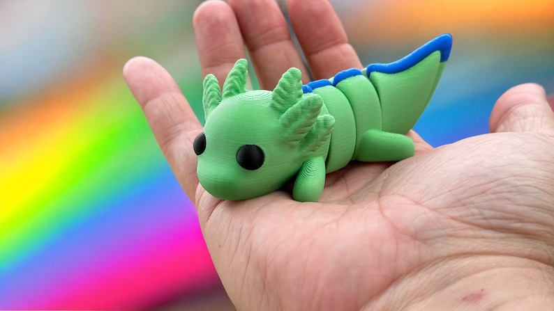 May include: A green and blue 3D printed axolotl figurine with black eyes. The figurine is posed with its head tilted to the side and its tail curled up.
