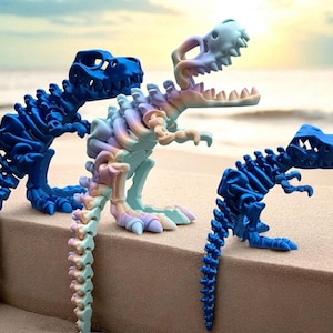 Puede incluir: Tres figuras de esqueletos de dinosaurios en tonos azules y pastel se exhiben sobre una superficie arenosa. Los dinosaurios est&aacute;n posados en diferentes posiciones, uno de ellos con un degradado arco&iacute;ris. El fondo muestra una escena de playa borrosa.