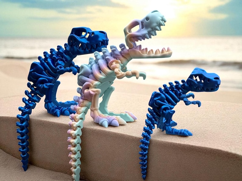 Puede incluir: Tres figuras de esqueletos de dinosaurios en tonos azules y pastel se exhiben sobre una superficie arenosa. Los dinosaurios est&aacute;n posados en diferentes posiciones, uno de ellos con un degradado arco&iacute;ris. El fondo muestra una escena de playa borrosa.